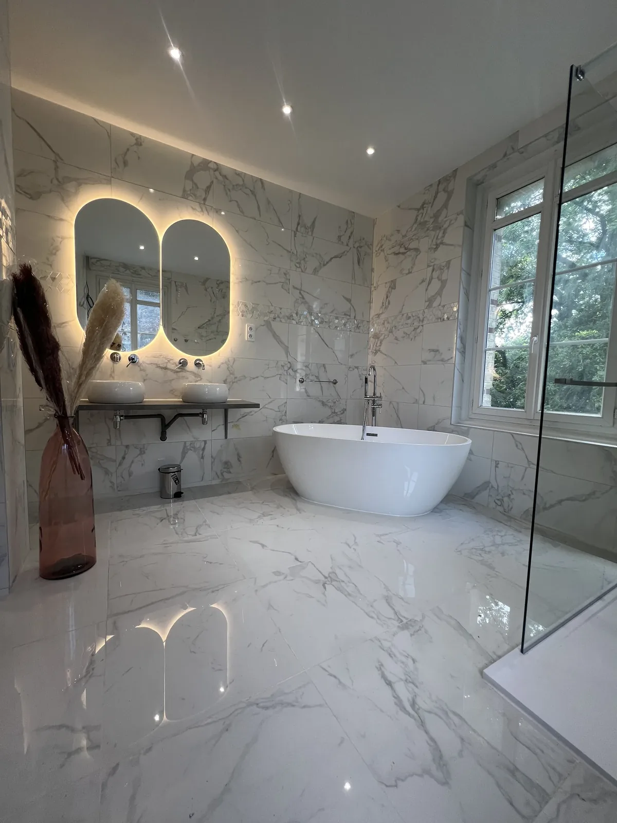 Salle de bain en carrelage effet marbre blanc, baignoire îlot et double miroir ovale rétroéclairé